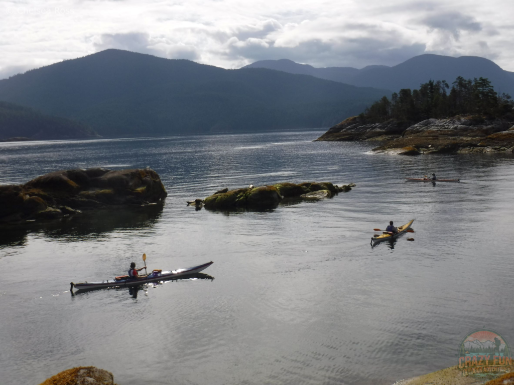 Three kayaks on the ocean leaving for the day kayaking in Desolation Sound. Rocks with algae are protruding out of the water with mountains in the backdrop.