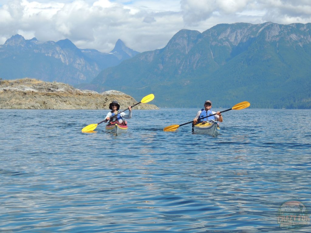 A lady kayaking in Desolation Sound in a red kayak to the left and a man is to the right of her kayaking in a yellow kayak with a brown rock behind the women and mountains in the backdrop.