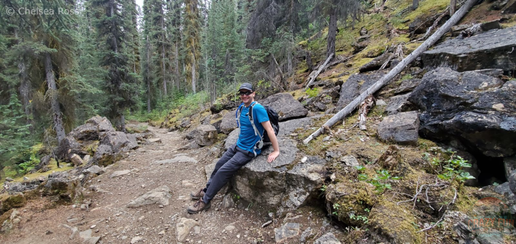 Man with blue t-shirt and backpack on his back is taking a break as he sits on a rock as he hikes up the steep part of the trail hiking to Laughing Falls. Rocks surround him as well as trees on both sides.