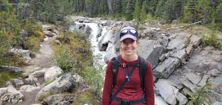 I'm standing in a red shirt in front of a small glacial grey and white waterfall as we make our way hiking to Laughing Falls. There was a small viewpoint to the right of the trail. There are grey rocks to the right of me and a small path with rocks to the left of me.