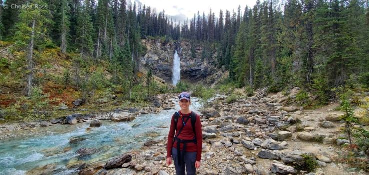 Lady in red shirt standing on rocks in front of the falls after hiking to Laughing Falls. The gorgeous blue glacial water in running to the left of me. There are trees on both sides of me.