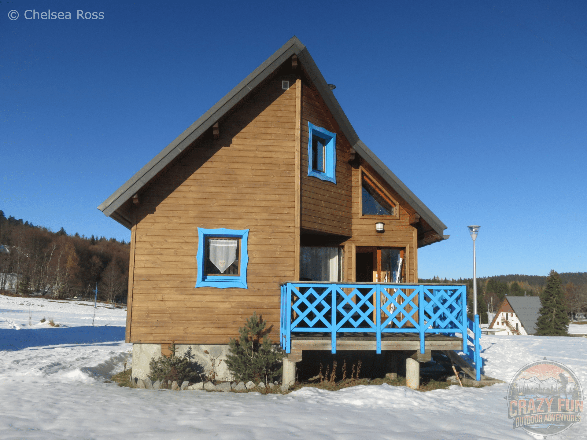 A quaint wooden chalet with blue window sills and blue railings on the patio. It's a gorgeous blue sunny day with snow lying on the ground.