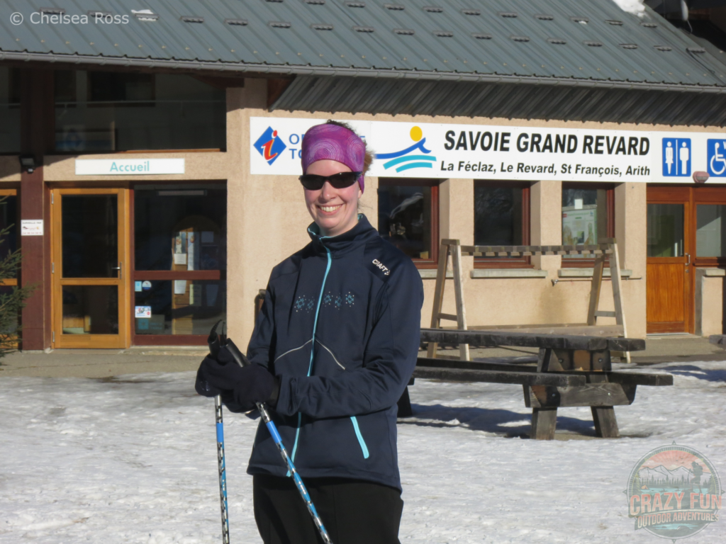 I'm standing with my purple tubular headwear and blue coat while holding my cross-country ski poles in front of the Tourism office in La Féclaz.