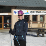 I'm standing in front of the tourism office in my blue coat with black pants and my tubular purple headwear while holding onto my ski poles. There's a sprinkling of snow on the ground for cross-country skiing in La Féclaz.