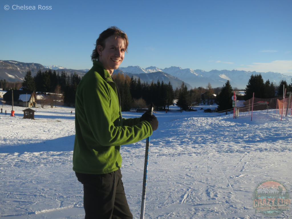 Scott is cross-country skiing La Féclaz while wearing his green hoodie and black pants. It's a gorgeous sunny day. Snow is covering the ground.