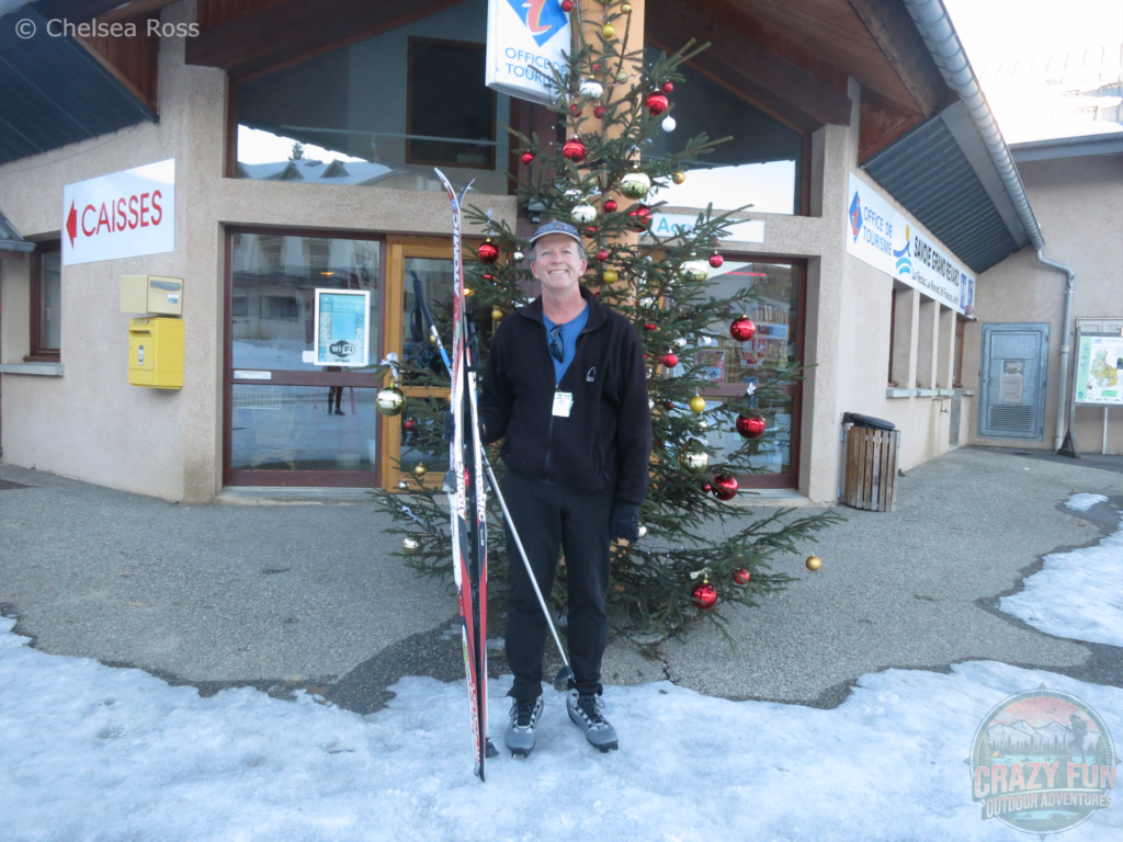 My dad standing with his skis to his right side in front of a Christmas tree at the tourism office. He's wearing black fleece and black pants.