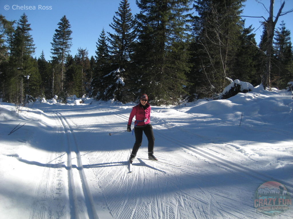 My mom trying out skate skiing in La Féclaz. She's in the middle of the classic cross-country skiing tracks wearing a pink coat and black pants. There's a nice ski base for cross-country skiing.