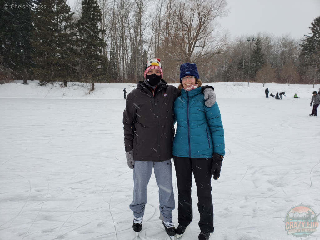 Kris and I standing on the ice with our skates on at Hawrelak Park. It's snowing outside as shown by white dots through the entire picture. The ice is also covered in snow. Kris (on the left) is wearing a penguin tuque with a face mask (for covid) and a black jacket, while I'm wearing a blue tuque with a teal jacket.