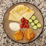White pita on the top of the plate with white yogourt dipping sauce below to the left, followed by tomatoes in the middle of the picture and sliced cucumbers to the right. Two salmon patties are on the bottom of the plate.