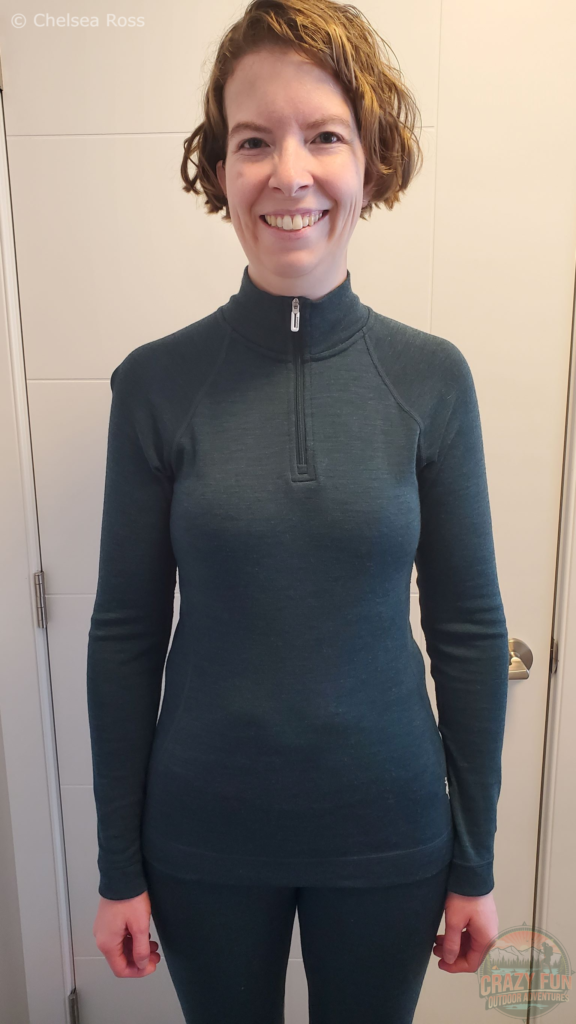 I'm standing in front of a white door while wearing my green Merino 250 Top Base Layer ready for cross-country skiing.