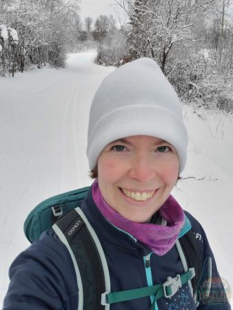 Keeping warm while wearing a Merino 250 Top Base Layer while cross-country skiing. She is wearing a green backpack, a white hat, purple tubular headwear and a blue jacket.