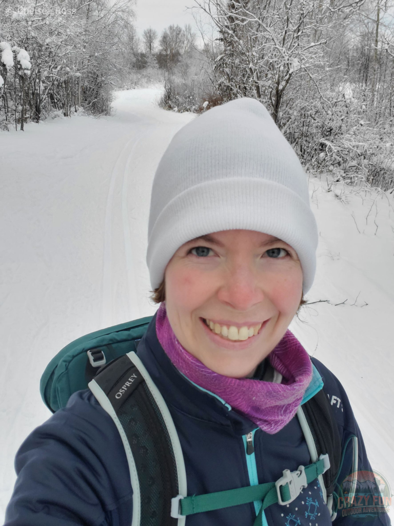 Keeping warm while wearing my Merino 250 Top Base Layer while cross-country skiing. I'm wearing a green backpack, a white hat, purple tubular headwear and a blue jacket.