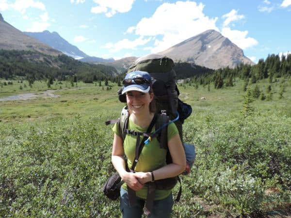 Lady smiling with a backpack in front of a mountain meadow in the background.