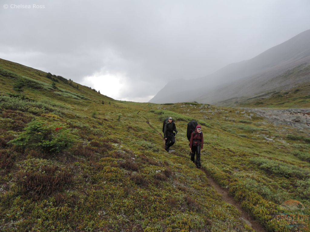My brother and I backpacking with our rain gear on us at the top of the pass after having passed through a rain storm. We are surround in green moss and a mountain to the right of us. 