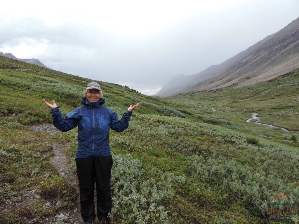 My mom in a happy state, the eternal optimist finds a way to stay positive backpacking in the rain. She's wearing a blue raincoat with black rain pants just after a rain storm standing in a meadow of moss at the top of a pass. 