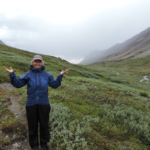 My mom in a happy state, the eternal optimist finds a way to stay positive backpacking in the rain. She's wearing a blue raincoat with black rain pants just after a rain storm standing in a meadow of moss at the top of a pass.