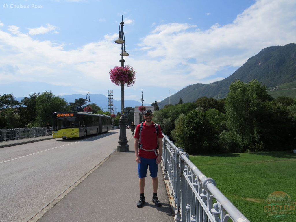 Kris with his orange shirt standing on a bridge in the middle of the picture with a bus saying Bolzano to the left of him.