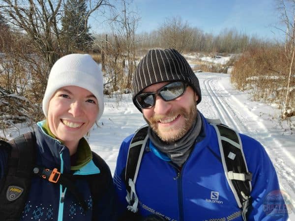 Couple cross-country skiing. Tracks and trees are behind them. 