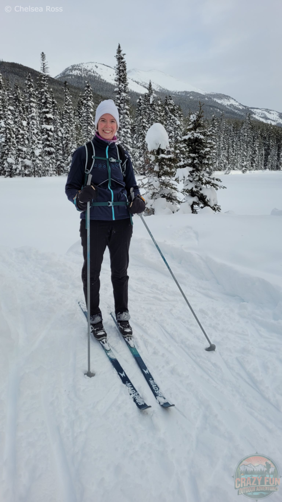 I'm standing on my cross-country skis wearing my Merino pant base layer underneath my black fleece pants.  It's keeping me warm on a cold winter day. I'm wearing a white hat with a blue coat and a green backpack. The trees and ground are covered in snow in the background.