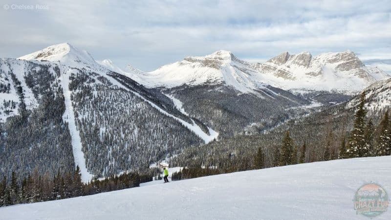 Gorgeous snowy view of the mountains across the valley. Lady is green coat skiing down the blue hill. 