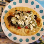 A gyro on a plate with feta, cucumbers, tomatoes, onion and sauce. The plate has blue dots around it. It's sitting on a placemat with a building behind it on the left and trees on the right.