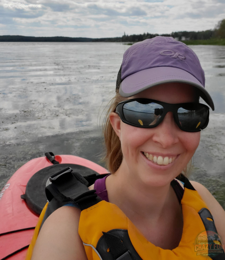 I'm sitting in a red kayak with my yellow life jacket, sunglasses and purple hat. It's a cloudy day on the water.