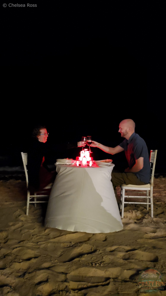 Me to the left and Kris to the right sitting on chairs for our romantic supper at the beach. Pitch black can be seen behind us. We are raising our glasses of wine and toasting.