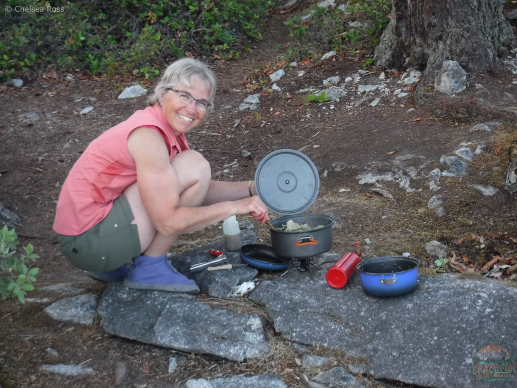 15 Awesome Kayaking Gifts for Moms: a pot is number five. My mom in a pink tank top and green shorts is crouched down mixing food in a pot cooking supper on the rocks.