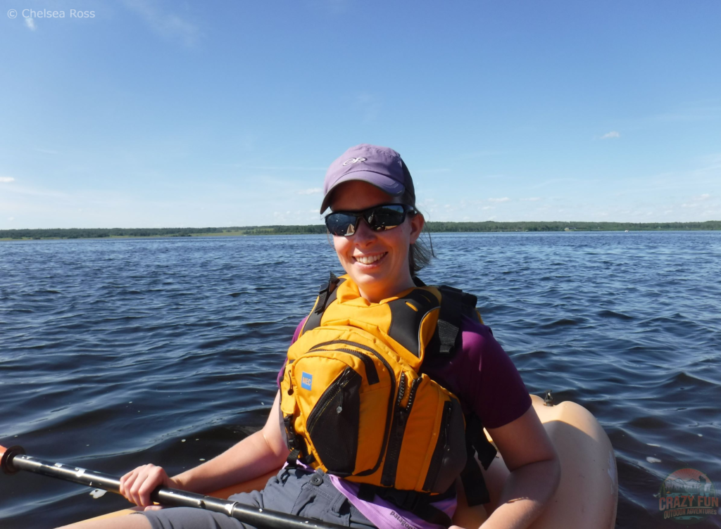 15 Awesome Kayaking Gifts for Moms: the first one is a life jacket. I'm sitting in a kayak with a purple shirt, yellow lifejacket, sunglasses and a purple hat. The lake can be seen behind me. 