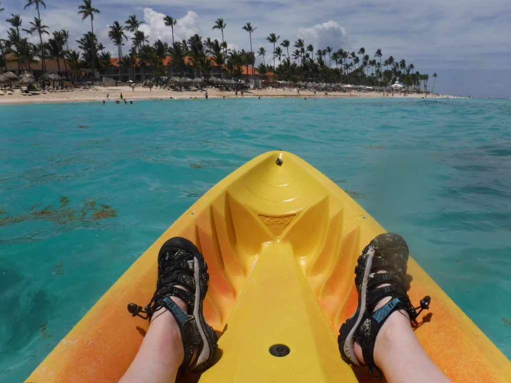 Taking a picture of my feet with sandals in the uncovered kayak. The ocean is seen in front with the beach and palm trees at the top of the picture.