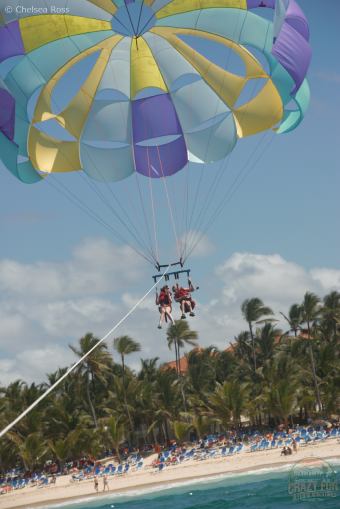 Top 10 Activities from Majestic Resorts: parasailing is number 5. Kris and I are in the air starting our parasailing trip. Palm trees and chairs filled with people are below. 