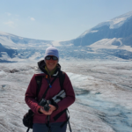 I'm standing in my purple jacket with my backpack on holding my camera in my hands. The glacier can be seen in the background.