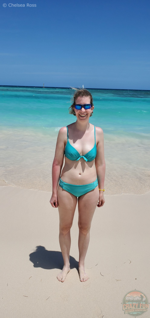 I'm wearing blue sunglasses while wearing my green bikini. I'm standing in front of the turquoise ocean. A sailboat can be seen on top of my head.