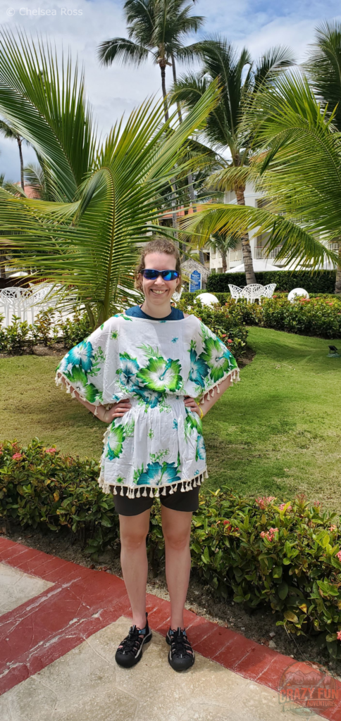 10 Tips to Avoid Sunburn: I'm wearing a shawl to cover my shoulders and back from the sun. The shawl is white with green and blue flowers. I have my hands on my hips to show it off. Green bushes and balm trees can be seen in the background. 