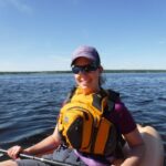 Lady wearing a bright yellow MEC Fulcrum Guide PFD while kayaking on a calm lake.
