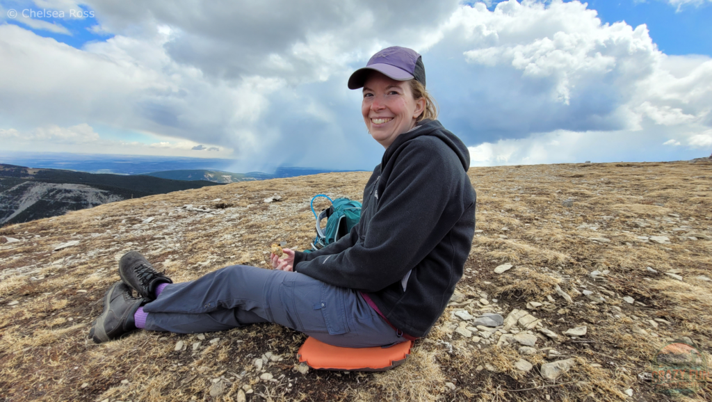 I'm sitting on a orange MEC seat cushion at the top of Prairie Mountain. Shale rocks are covering the mountain top. Clouds are starting to roll in. 