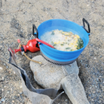 The GSI Escape 2L Pot in blue cooking rice, chicken, green beans, and corn for supper. The pot is sitting on the MSR rock that is held up by a rock on the sand. The MSR bottle sits behind the pot.