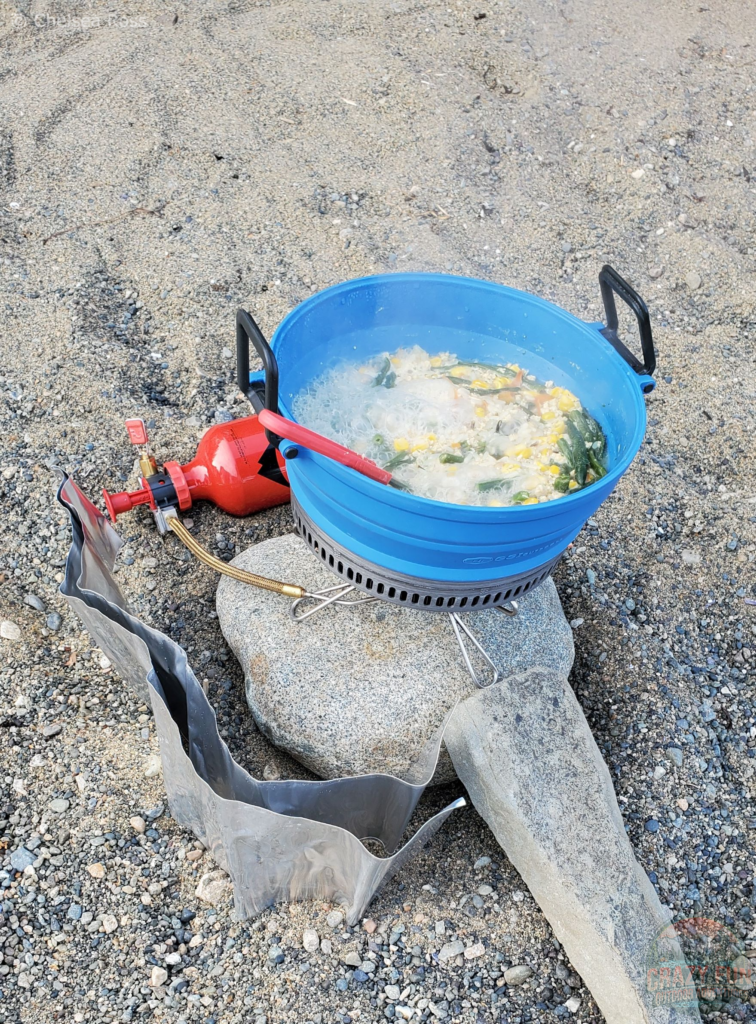 The GSI Escape 2L Pot in blue cooking rice, chicken, green beans, and corn for supper. The pot is sitting on the MSR rock that is held up by a rock on the sand. The MSR bottle sits behind the pot. 