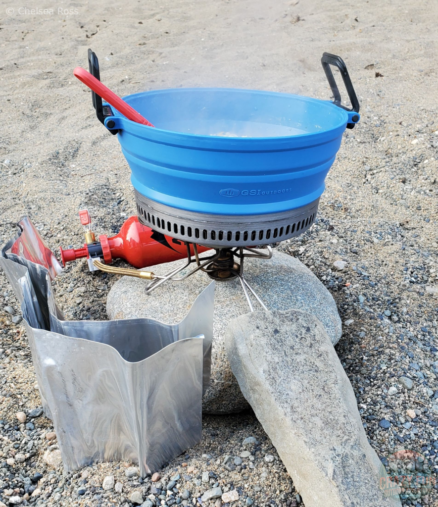 The GSI Escape 2L Pot in blue on the MSR stove sitting on a rock on the beach. The pot that has supper in it can be seen in full height with black handles on the sides and a red spoon in the pot. 