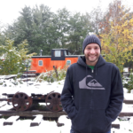Kris standing in his black fleece in front of an orange train with snow on the ground.