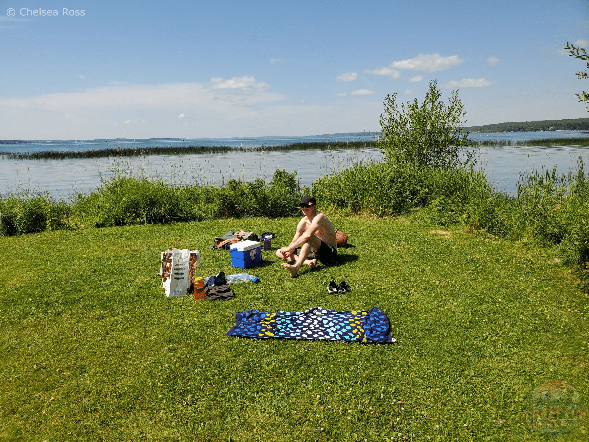 Scott in his swimsuit on the grass with a towel, cooler, food in front of Sylvan Lake.