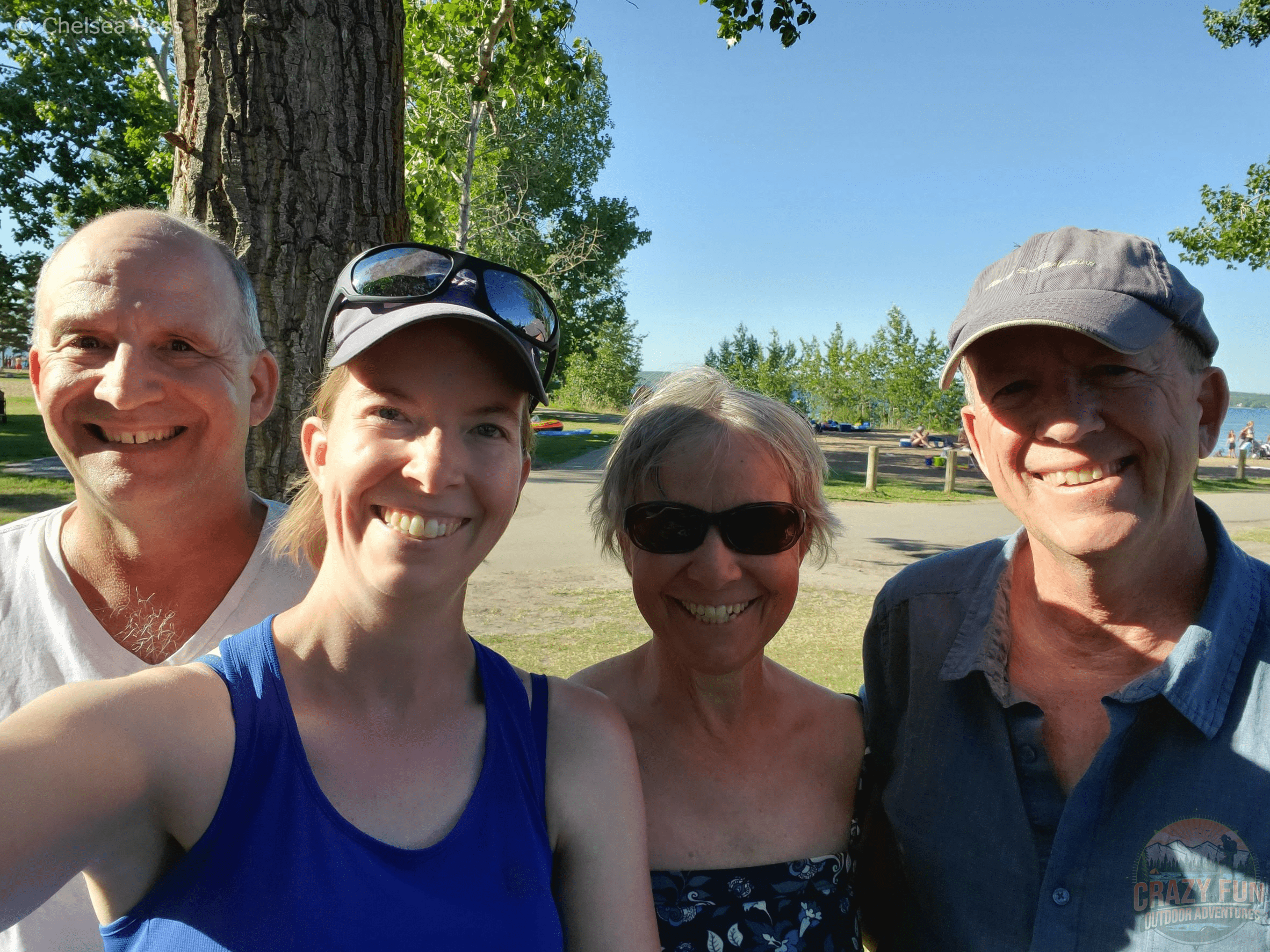 Selfie of four people. Left to right: white shirt, blue tank top, blue swimsuit and blue shirt in front of trees.