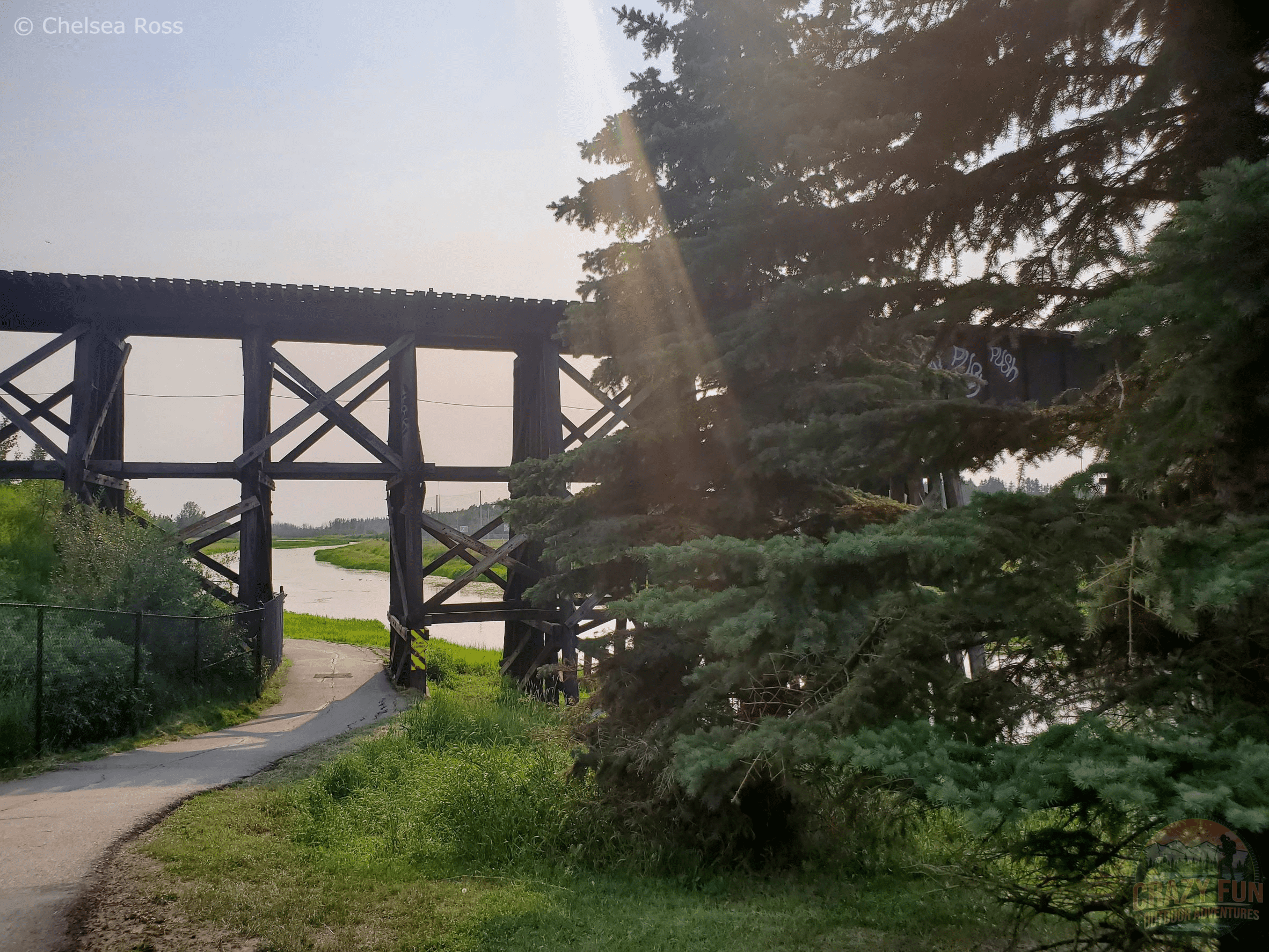 A railway bridge on top of the pathway with the river to the right side with grass and trees on the right side. 