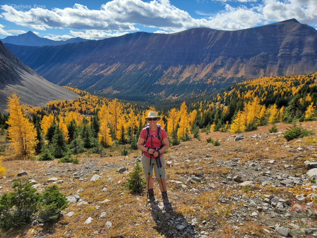 The best larch hikes showcasing my mom at Little Arethusa with beautiful larches and a mountain behind her.