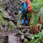 I'm standing on rocks on the WCT with my blue raincoat, black rain pants, and blue and black gaiters while wearing a backpack. Our WCT packing list will help to pack for a wonderful backpacking trip.