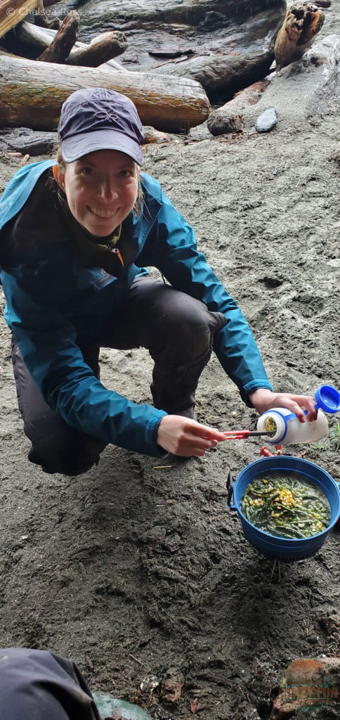 I'm squatting with a blue jacket on the beach while cooking my dehydrated meal in a pot over a stove. I'm pouring the contents of my water bottle (dehydrated food) into the pot.