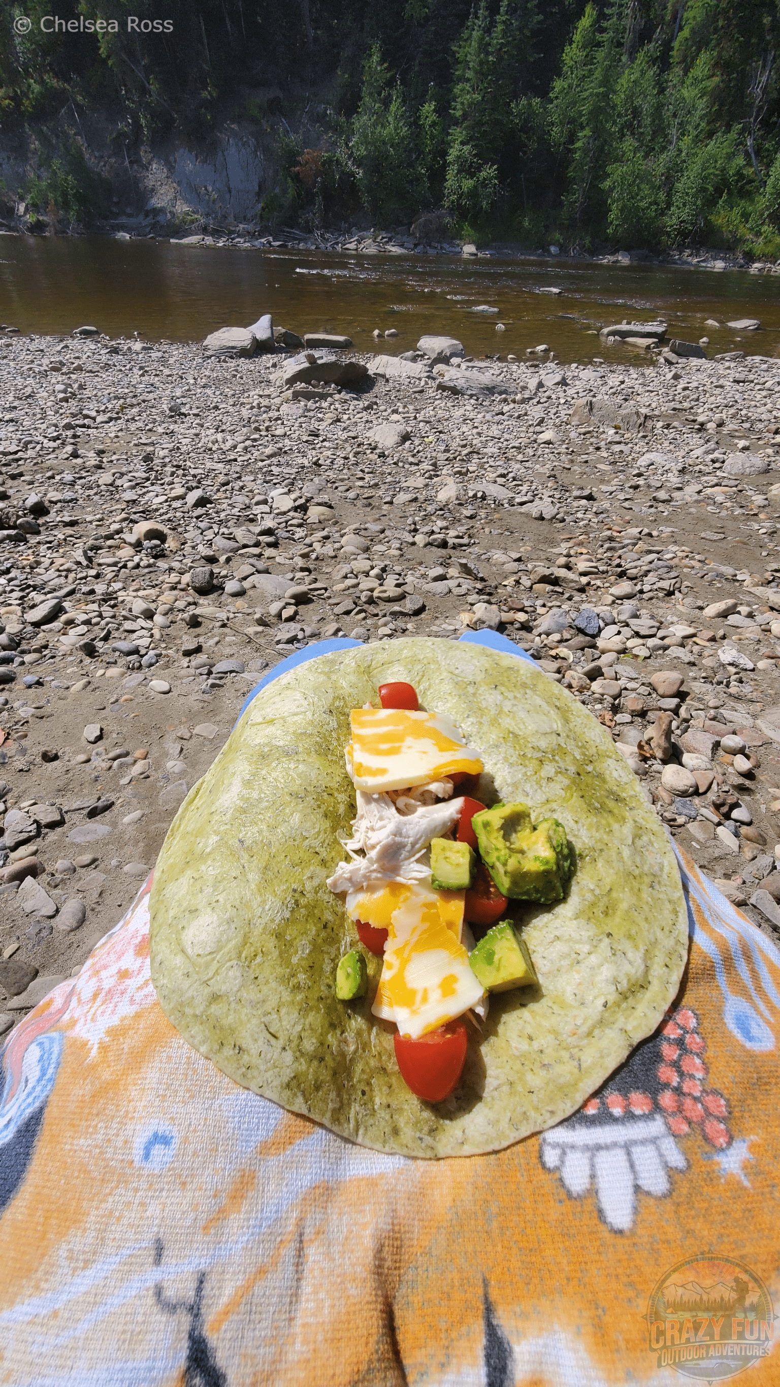 A cheese, avocado, chicken and tomato wrap on my towel covering my legs with the river in the background.