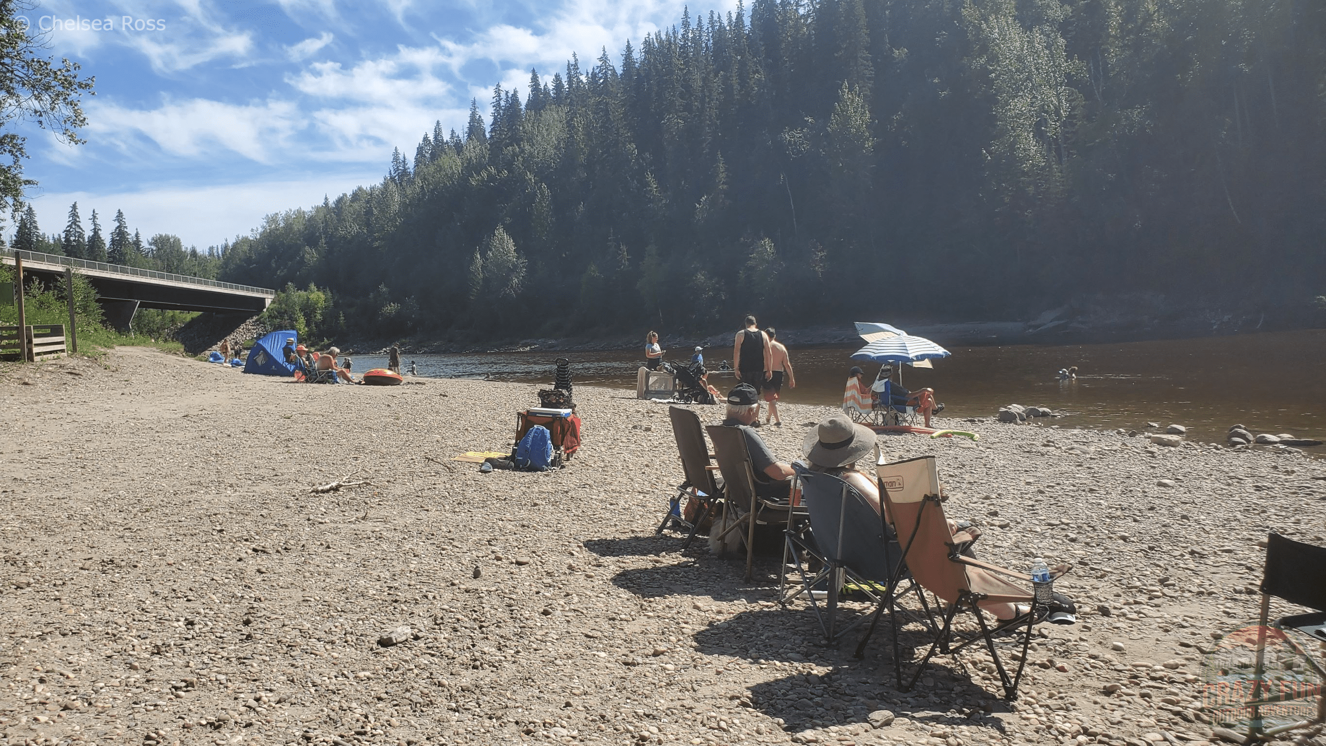 Chairs and people sitting on the rocky beach in front of the water. A bridge is on the left side and trees in the background.