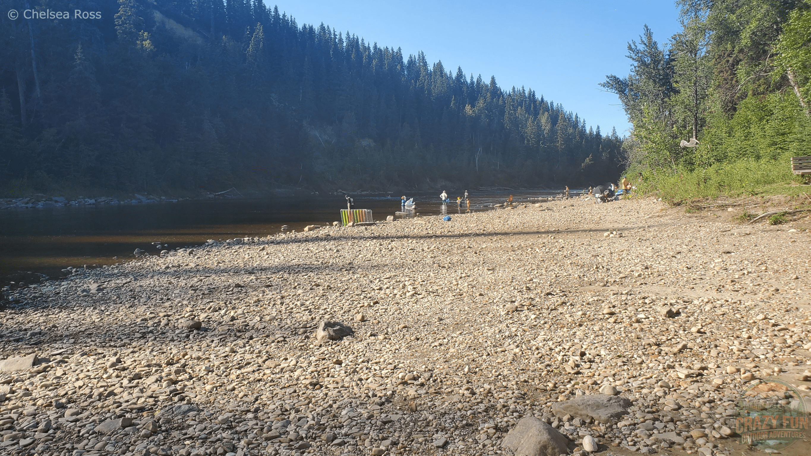 People can be seen throughout the rocky beach with the river and trees in the background.