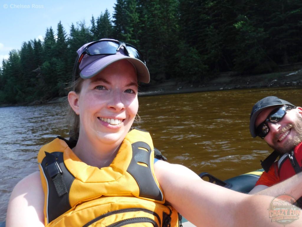 Taking a selfie of Kris and I in the raft on the water. We're both wearing hats and our PFDs on our Pembina River rafting experience. The water is calm behind us.